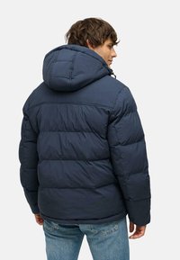 Giacca puffer blu navy con cappuccio, caratterizzata da sezioni trapuntate e una vestibilità flessibile. Abbinata a jeans azzurri chiari.