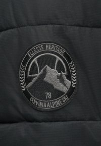 Μαύρο ύφασμα με κεντητό βουνό, αριθμό 78, κυκλικό λογότυπο και κείμενο "ELLESSE HERITAGE" και "CERVINIA ALPINE SKI."