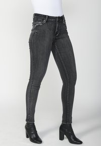 Črne ozke kavbojke z zbledelim videzom, opremljene z dekorativnimi kovinskimi elementi na žepih in klasičnim petim žepnim dizajnom. Nosi se z gležnjarji.