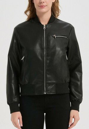 Femme portant une veste bomber en cuir noir avec des poches à fermeture éclair sur la poitrine et les côtés, des poignets et une ceinture côtelés, debout devant un fond uni.