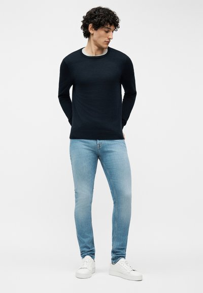 Άνδρας με σγουρά μαλλιά, φορώνοντας μαύρο πουλόβερ με μακριά μανίκια, στενά ανοιχτό μπλε τζιν και λευκά αθλητικά παπούτσια, στέκεται με τα χέρια πίσω από την πλάτη.