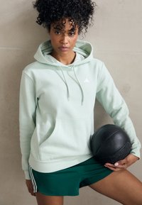 Sweatshirt capuz verde-claro com bolso frontal e logótipo branco, combinado com calções verde-escuro, segurando uma bola de medicina preta.