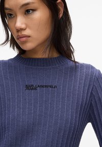 Pleteni pulover v temno modri barvi z rebrasto teksturo, s krožnim ovratnikom in črnimi vezenimi logotipom "KARL LAGERFELD JEANS" na prsih.