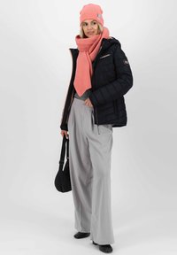 Schwarze Daunenjacke, grauer Strickpullover, pinker Schal und Mütze; weite graue Hose, schwarze Tasche und schlichte schwarze Schuhe. Weiche Texturen sichtbar.