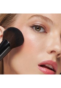 Make-upkwast met een dichte, zwarte harenkop die blush aanbrengt op een lichte huid. De oogmake-up is subtiel met een natuurlijke lipkleur.