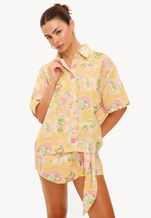 Chemise à manches courtes à boutons en tissu jaune avec un motif de fruits en rose et vert. Accompagnée d'un short assorti avec un détail de lien.