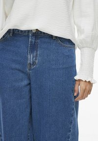 Zilās denim bikses ar taisnu kāju griezumu, aprīkotas ar priekšējo pogu un rāvējslēdzēju. Baltā blūze ar teksturētu audumu un krokām pie piedurknēm.