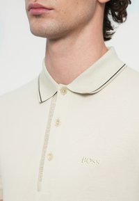 Béžové polo tričko s textúrovanou tkaninou, čiernym lemovaním goliera a tromi gombíkmi. Na hrudi má vyšívané logo "BOSS".