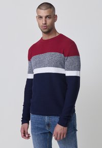 Maglione lavorato a maglia con una sezione superiore rossa, una parte centrale grigia, una striscia orizzontale bianca e una parte inferiore blu navy. Texture a coste, collo rotondo.