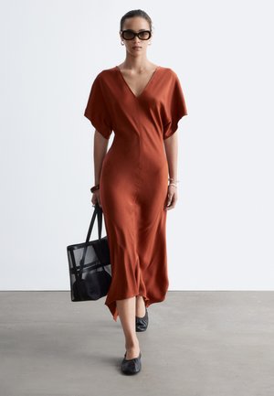 Femme marchant à l'intérieur portant une robe midi couleur rouille, des chaussures plates noires, des lunettes de soleil foncées, portant un sac fourre-tout noir.