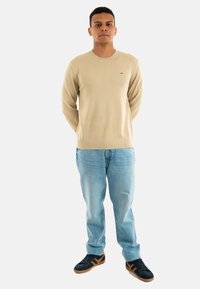 Maglione beige in maglia con scollo rotondo, abbinato a jeans azzurri chiari e sneaker scure con strisce arancioni. Design semplice.