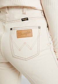 Vaqueros de mezclilla blancos con una etiqueta de cuero Wrangler en el bolsillo trasero, con costuras en contraste y un diseño clásico de bolsillo.