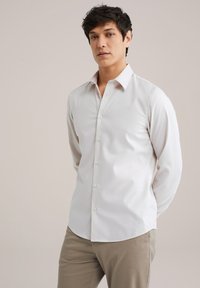 Camisa blanca de manga larga con cuello abotonado, con una textura suave y dobladillo recto, combinada con pantalones beige.
