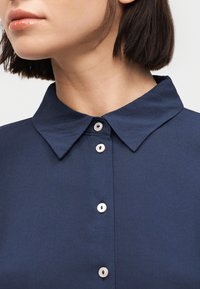 Γυναίκα με κοντά σκούρα μαλλιά, φορώντας μπλε σκούρο πουκάμισο με γιακά, κουμπωμένο μέχρι τον λαιμό, σε απλό λευκό φόντο.