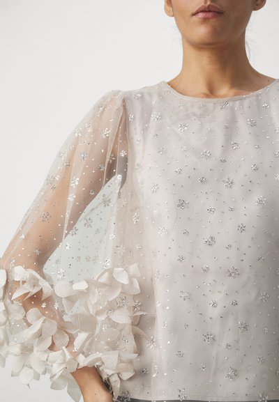 Sheer beige blouse featuring floral embellishments at the sleeves, accented with silver sequins and a subtle textured fabric.