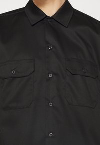 Chemise noire boutonnée avec deux poches à rabat devant, portée par une personne visible du cou jusqu'au milieu de la poitrine.