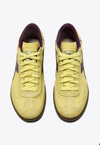Sneaker in camoscio giallo con una striscia laterale viola, lacci testurizzati e suola in gomma marrone. La vista dall'alto mette in evidenza linee pulite e un design semplice.