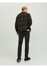 Camicia flanella con motivo a quadri neri e dettagli marroni, indossata sopra una maglietta verde scuro, abbinata a jeans neri e scarpe nere, vista da dietro.
