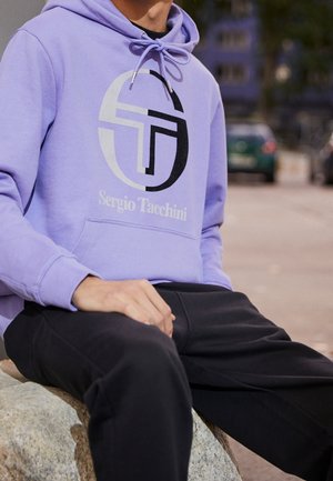 Felpa di cotone viola con un grande logo bianco e nero e il testo "Sergio Tacchini". Abbinata a pantaloni di felpa neri, seduta su una pietra.