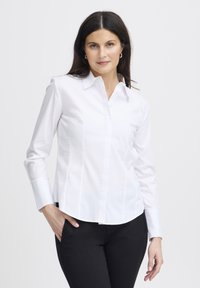 Camicia bianca con maniche lunghe, dotata di colletto classico, cuciture strutturate e orlo curvo. Abbinata a pantaloni neri.