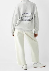 Grauer Pullover mit hohem Kragen, marineblauer Schrift auf der Rückseite und elfenbeinfarbene Hosen mit weitem Bein. Die Schuhe sind weiße Sneakers mit dezenten Details.