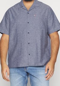 Camicia a maniche corte con bottoni in tessuto blu scuro a trama, dotata di tasca sul petto e patch con logo. Abbinata a jeans azzurri.
