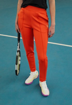 Osoba stojąca na niebieskim korcie tenisowym trzyma czarno-białą rakietę tenisową, ubrana w jaskrawo pomarańczowe spodnie i białe tenisówki z fioletową podeszwą.