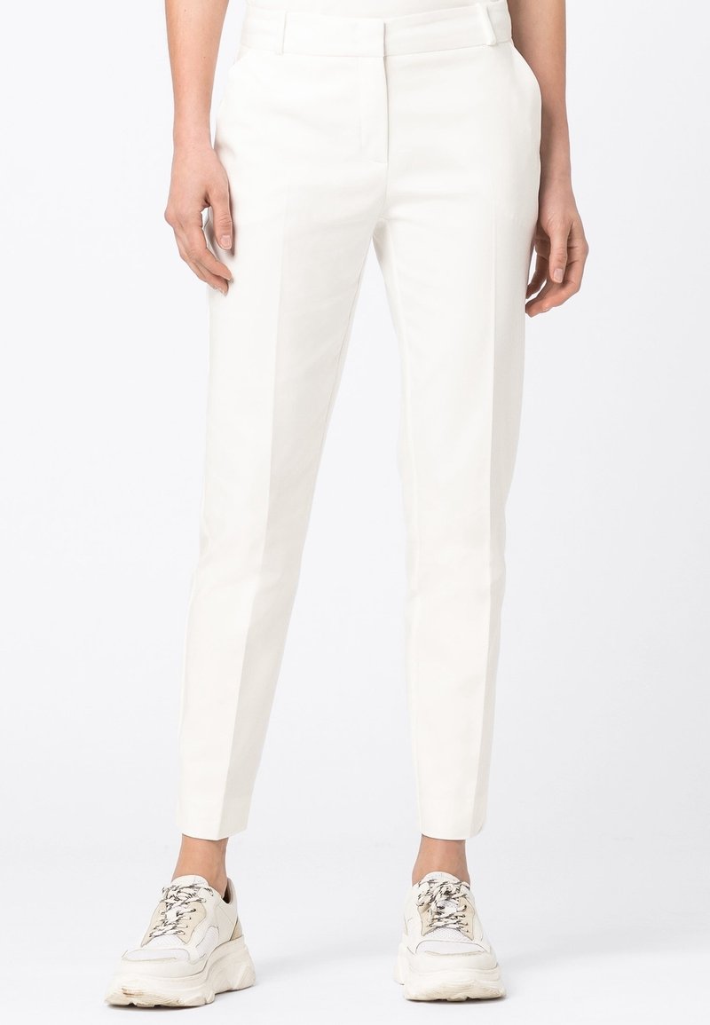 Pantaloni bianchi su misura indossati con sneakers di colore chiaro, che mettono in risalto uno stile moderno e informale su uno sfondo chiaro e semplice.