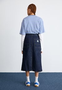 Gonna di jeans con due grandi tasche posteriori, camicia a righe blu e bianche a maniche corte indossata sopra una camicia bianca a maniche lunghe, acconciatura a treccia.