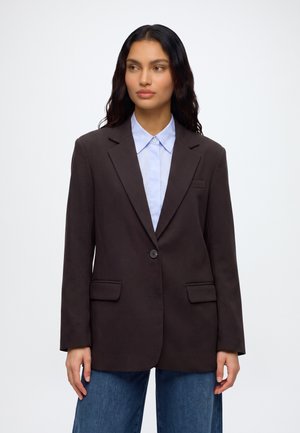 Femme aux longs cheveux foncés portant un blazer marron foncé par-dessus une chemise bleu clair à col et un jean bleu sur un fond uni.