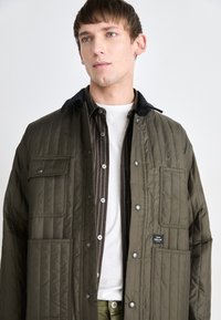 Olivgrüne gesteppte Jacke mit einem schwarzen Cordkragen, großen Vordertaschen und Druckknöpfen, geschichtet über einem gestreiften Hemd und einem weißen T-Shirt.