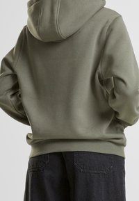Sweat à capuche vert olive, en tissu lisse. Présente des poignets et un ourlet côtelés, avec une coupe décontractée et des coutures visibles.