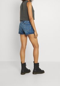 Persona vestida con una camiseta sin mangas oscura, pantalones cortos de mezclilla azules y botas negras con cordones, de pie contra un fondo claro y liso.