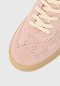 Zapatilla deportiva de ante rosa con cordones crema, presenta detalles de costura y suela de goma color chicle. Textura suave que contrasta con la parte superior blanda.