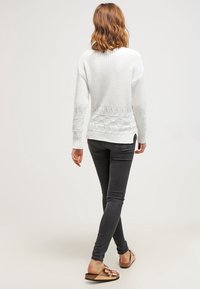 Biały sweter dzianinowy z teksturowanym wzorem na dole, zestawiony z czarnymi obcisłymi dżinsami i jasnobrązowymi sandałami. Modelka pokazana od tyłu.