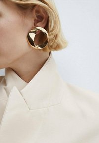 Mujer rubia que lleva un pendiente de aro dorado grande, brillante e irregular y una chaqueta crema de cuello alto, con un fondo blanco.