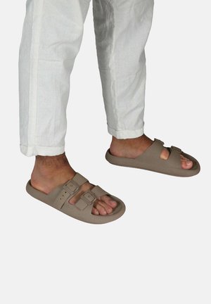 Sandales brunes à enfiler avec deux sangles réglables, surface texturée et design ouvert à l'avant, portées avec un pantalon en lin blanc ample.