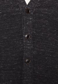 Tmavě šedý cardigan s texturovaným pleteným vzorem, s výstřihem do V, pěti černými knoflíky a pod ním viditelným bílým tričkem.