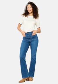 Jeans de mezclilla azul acampanados con dos bolsillos delanteros de parche, cintura alta y detalles sutiles de costura, combinados con una camisa crema con ribetes negros.