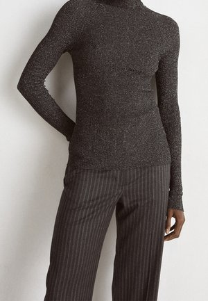 Gros plan sur une personne portant un pull à col roulé ajusté à manches longues, sombre et pailleté, ainsi qu'un pantalon ample sombre à fines rayures.