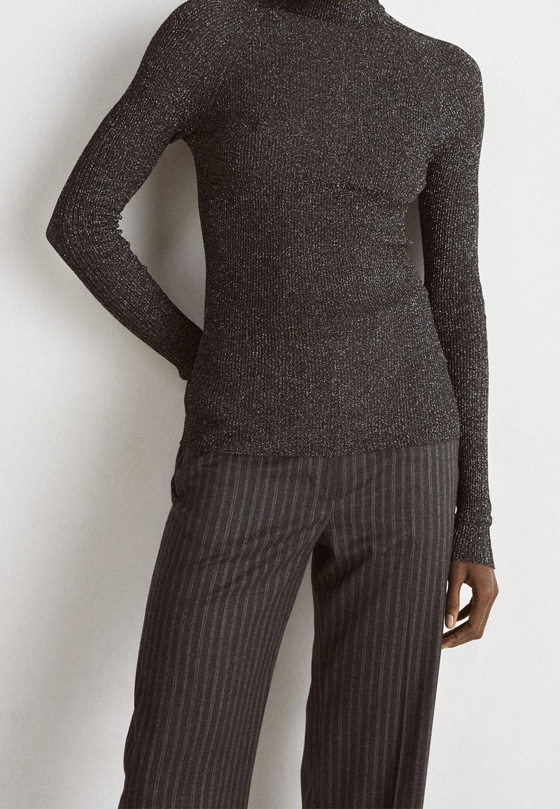 Gros plan sur une personne portant un pull à col roulé ajusté à manches longues, sombre et pailleté, ainsi qu'un pantalon ample sombre à fines rayures.