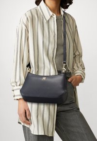 Borsa a tracolla in pelle blu navy con una forma strutturata, logo in metallo dorato e tracolla regolabile; abbinata a una camicia a righe e jeans grigi.