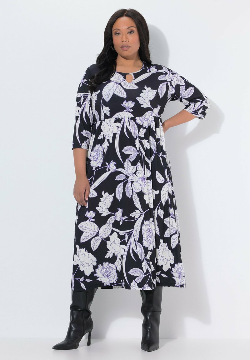 Vestido largo negro con estampado floral, mangas tres cuartos, con diseños florales en blanco y lavanda, y escote con abertura en forma de gota. Combinado con botas negras hasta la rodilla.