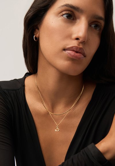 Γυναίκα που φοράει μαύρο τοπ, χρυσό κρίκο σκουλαρίκι και στρωμένα χρυσά κολιέ, ένα με μενταγιόν "S", κοιτάζει ελαφρώς στο πλάι.