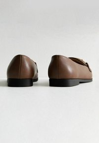 Braune Lederloafers mit runder Zehe, minimaler Details und einer flachen schwarzen Sohle, von hinten betrachtet. Glatte Textur und einfaches Design.