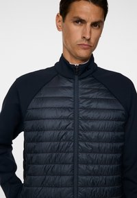 En man med ett allvarligt uttryck bär en marinblå zip-up jacka med ett kviltat framstycke och släta ärmar, mot en enkel vit bakgrund.