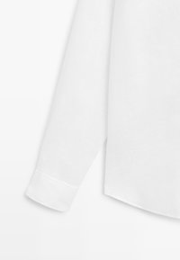 Chemise blanche à manches longues avec un poignet visible et un ourlet droit sur un fond uni.