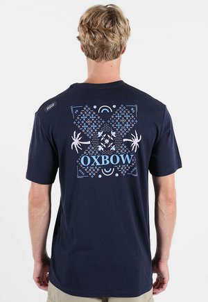 Homme aux cheveux blonds courts portant un T-shirt bleu marine avec un motif géométrique et floral décoratif bleu et blanc, et le texte "OXBOW" au dos.