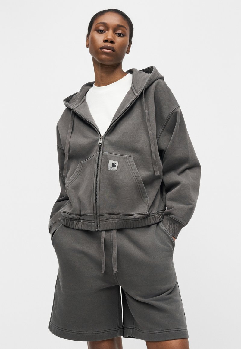 Giovane che indossa una felpa con cappuccio grigio scuro con zip e tasche frontali e pantaloncini coordinati fino al ginocchio, in piedi con le mani nelle tasche.