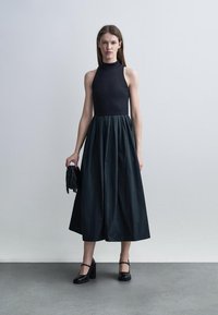 Kobieta stoi przy gładkiej ścianie, ubrana w czarną sukienkę bez rękawów, trzyma małą czarną torebkę z frędzlami i nosi czarne buty na obcasie typu Mary Jane.